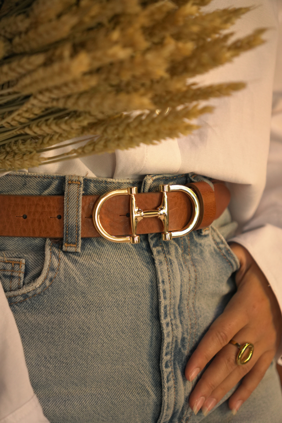 Détail de la boucle dorée en forme de mors de la ceinture Jeanne Atelier Nappa en cuir camel, sur un jean clair