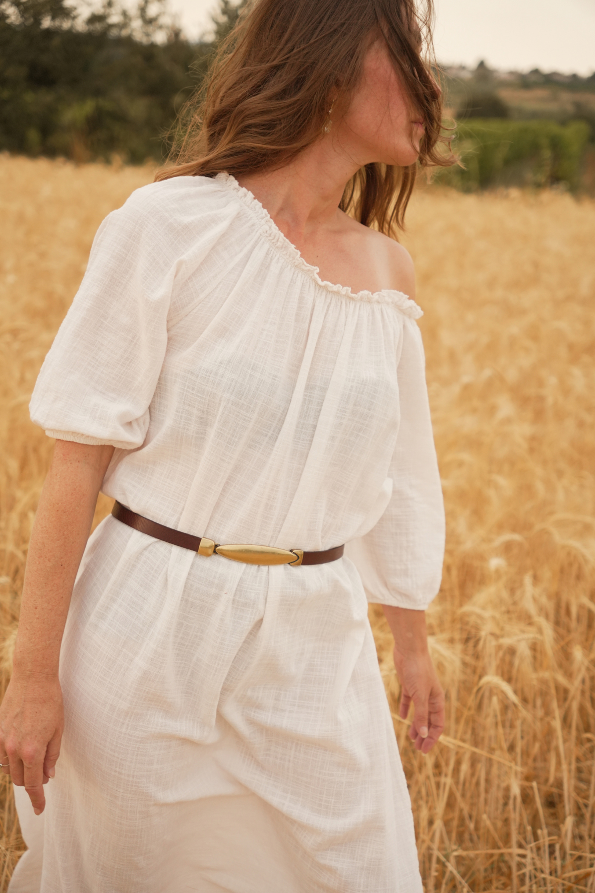 Ceinture fine Rachel Atelier Nappa en cuir marron à boucle ovale dorée, portée à la taille sur une robe blanche dans un champ de blé