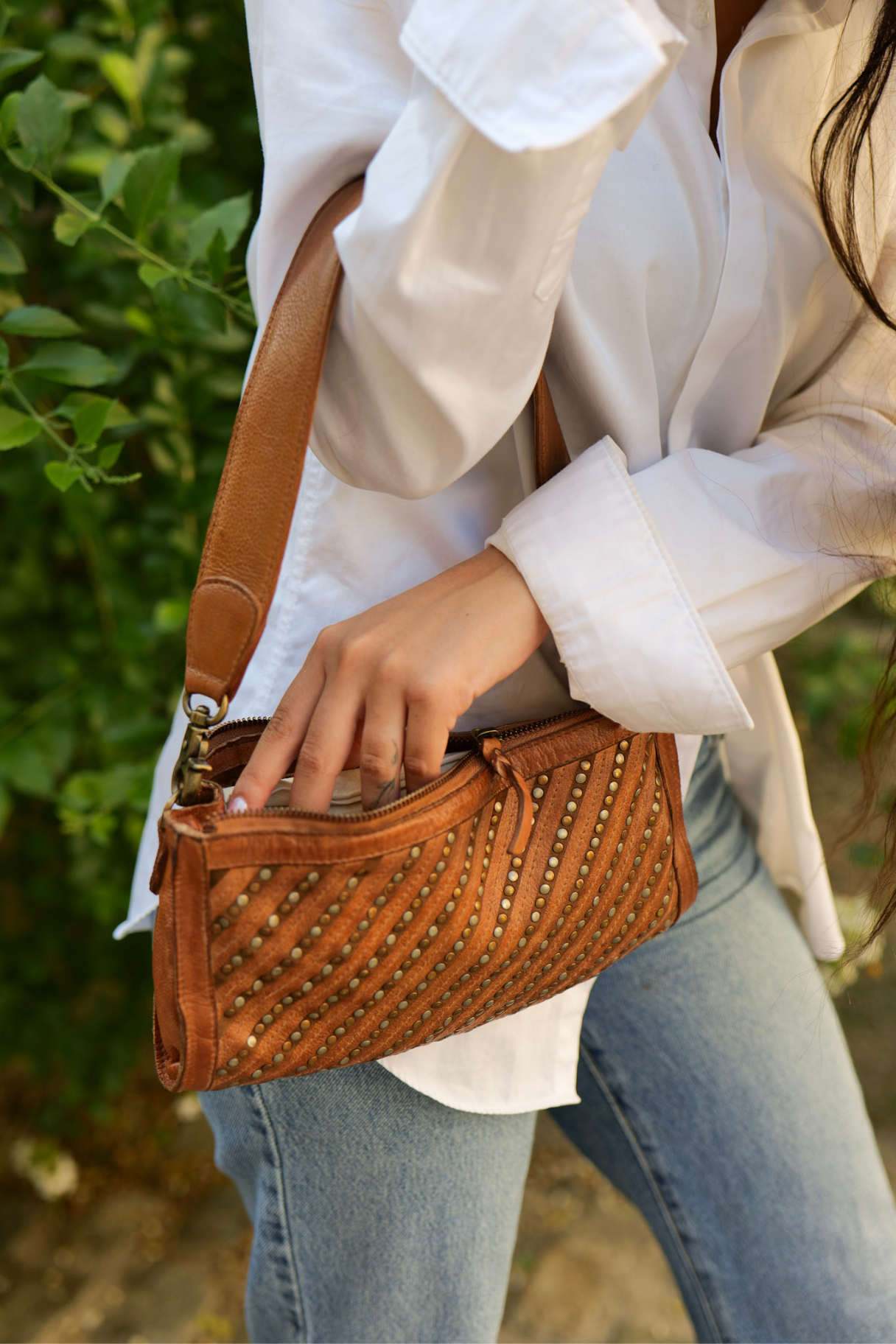 Gros plan sur pochette bandoulière en cuir cognac avec détail des clous métalliques et fermeture zippée