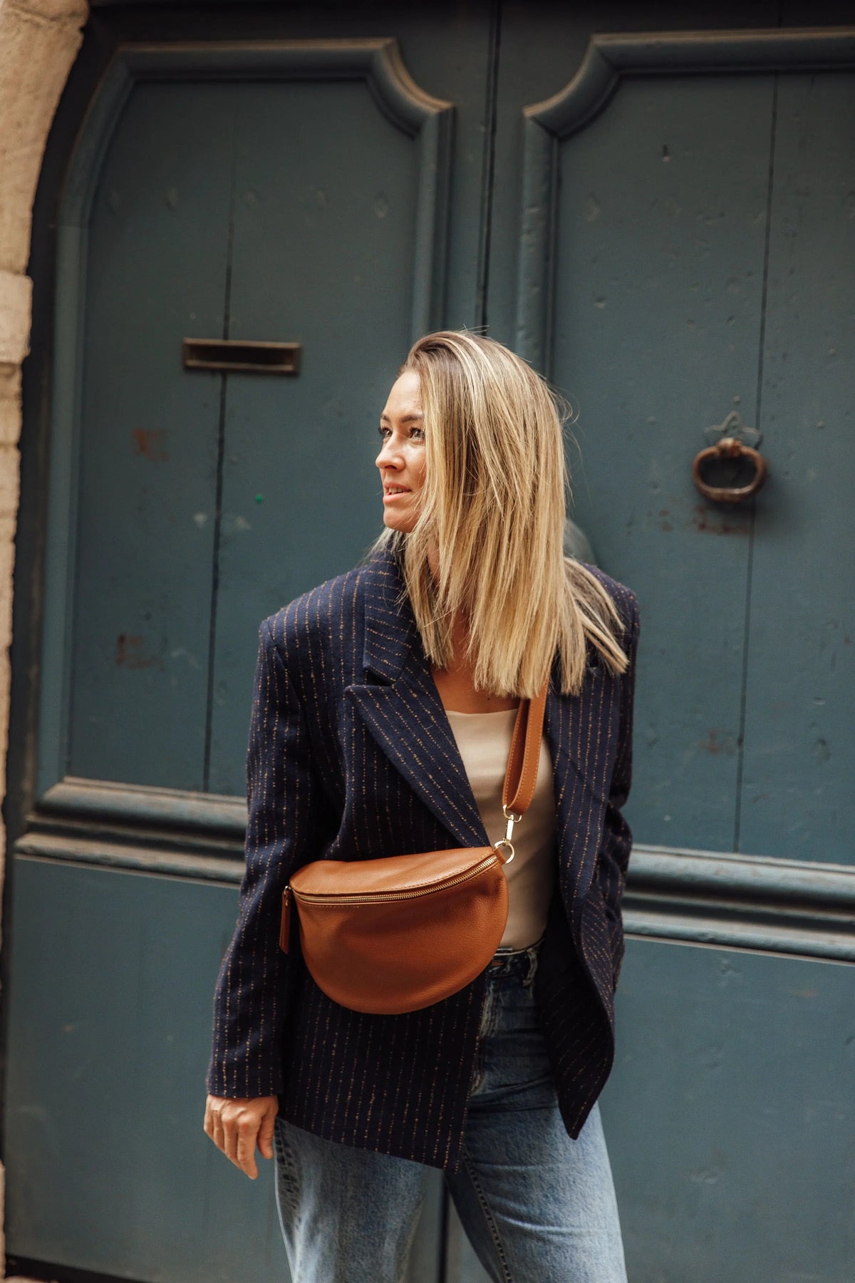 Sac banane Jeanne en cuir grainé camel porté en travers sur un pull beige et un jean